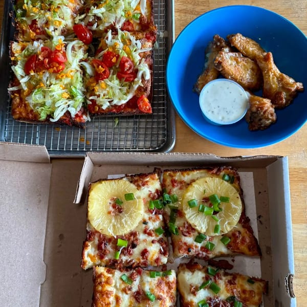 Two Sodough Square pizzas, one with lettuce and tomatoes, another with pineapple, alongside chicken wings.