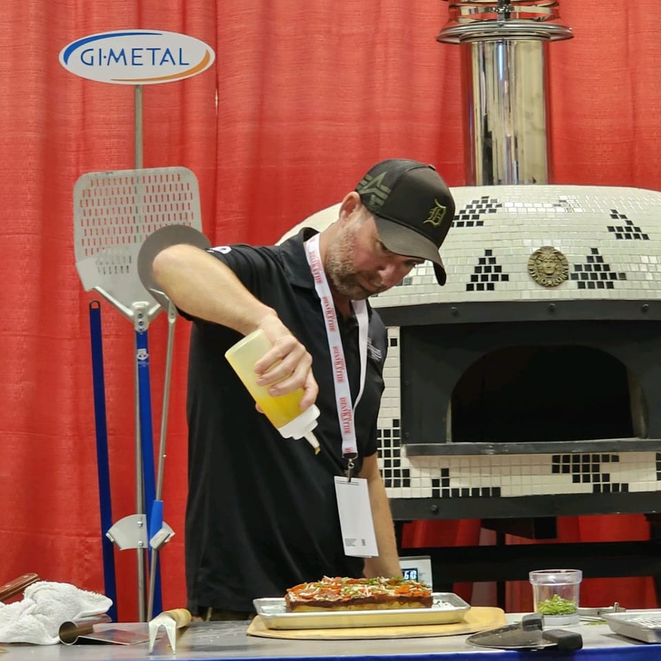 A chef drizzles oil on a rectangular pizza with pepperoni and vegetables, next to a pizza oven.