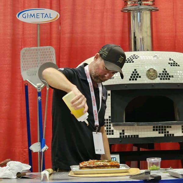 A chef drizzles oil on a rectangular pizza in front of a tiled pizza oven.