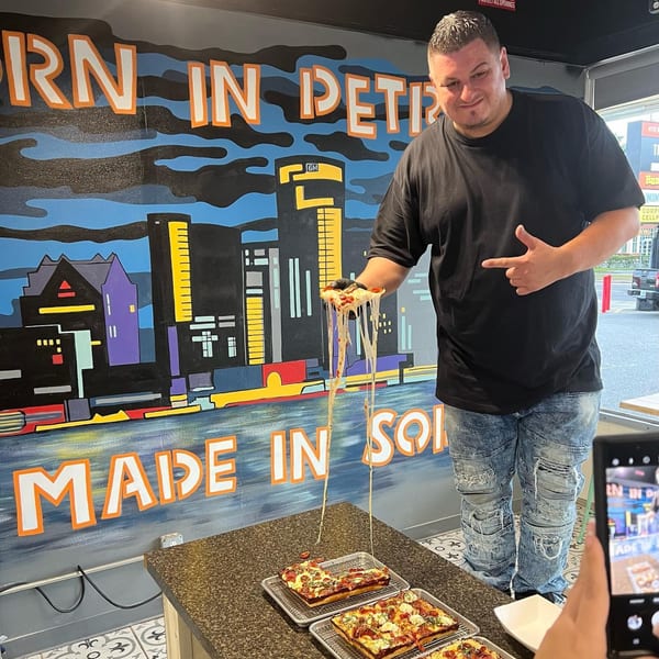 A man holds a slice of Sodough Square pizza with long cheese pulls, in front of a city mural.